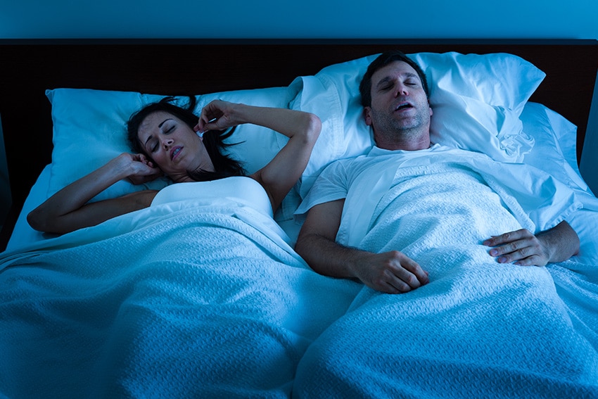 man snoring in bed while his wife plugs her ears shut